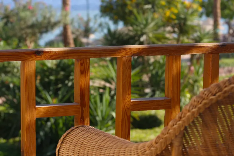 Holzgeländer und Korbsessel auf einer Terrasse mit Blick auf einen sonnigen Garten